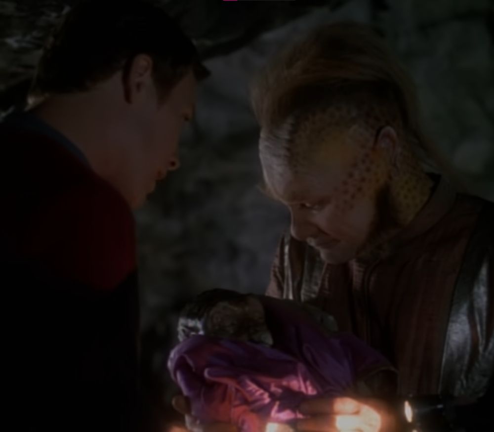 2 men holding a baby that looks like the eraserhead baby, it is swaddled in a coat. one man is an alien, the other isn't an alien, he's just an asshole. 