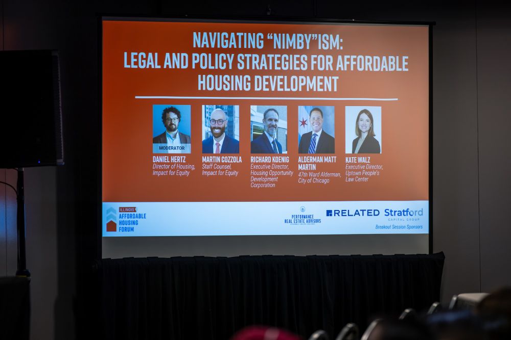 A powerpoint slide on a big screen in an auditorium with a red backer has a white title, "Navigating "NIMBY"ISM: Legal and Policy Strategies for Affordable Housing Development." There are five photos in a row beneath the title, however they are not super visible.