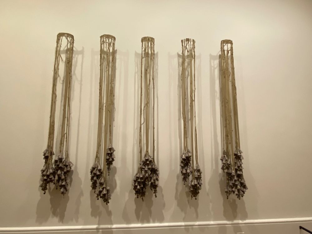 An installation of five macrame hangers; approximately five to six feet in height. Each hanger ends with ornamentation made from notes from community members to ancestors, shredded African lost-wax filigree brass trade beads, vintage African lost-wax brass bell beads, faux suede cord, paper pulp, white clay, and glue.