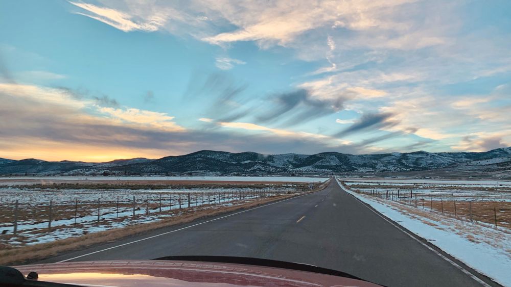 The sun is starting to set behind the thick layer of clouds on the horizon near Koosharem, Utah, creating a golden glow above the mountain line, with some greyish blue spots where the clouds are too thick for the light to pass through. Further up in the sky are some wispy clouds steaming across the sky, echoing the same colors as the horizon line. All across the ground and mountain and a thin layer of melting snow.