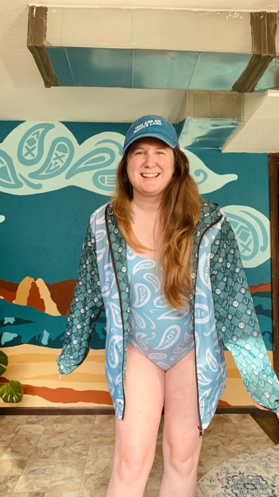 Artist, Ali Harris, is wearing a one-piece swimsuit and matching windbreaker that feature blue paisley bandana print she designed—the print is a modern, oversized take on the classic bandana paisley. The sleeves of the jacket have a green longhorn skull checkered print, also designed by Ali. On the wall behind her is a mural she painted of the Kolob Canyons 5 fingers from Zion National Park—the clouds in the mural feature the same paisley swirls in a much larger size.