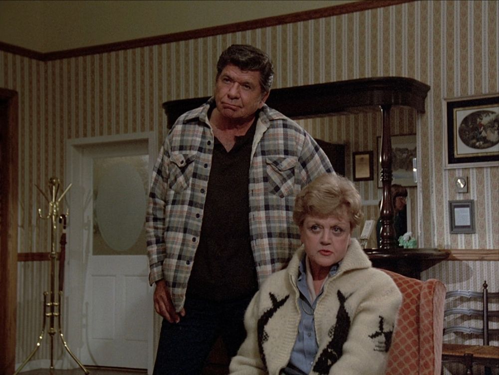 Claude Akins as Captain Ethan Cragg (left) and Angela Lansbury as Jessica Fletcher (right) in MURDER, SHE WROTE season 1. She is wearing a cream cardigan with black fish stitched into the sides and the left seat. Ethan is standing while Jessica is sitting.