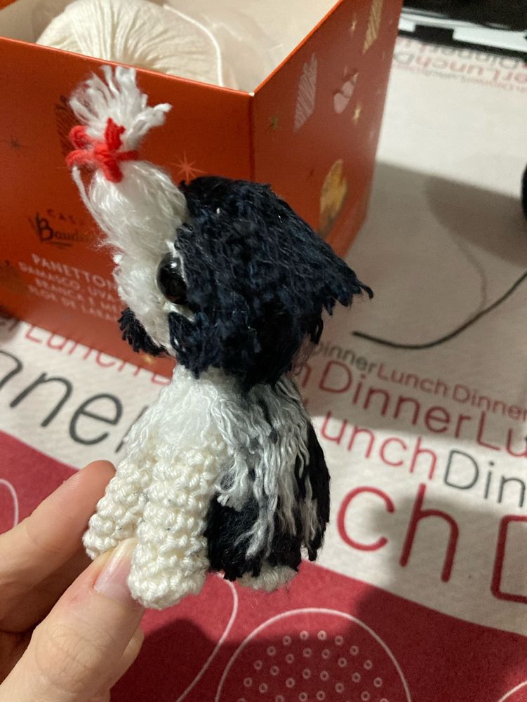 Cachorrinho shitzu feito de crochê preto e branco e com um lacinho vermelho nos pelinhos da cabeça. Fiz um pelo longo nas costas, no peito e na cabeça, já as patas da frente ficaram apenas com os pontos do croche mesmo. O boneco está de lado.