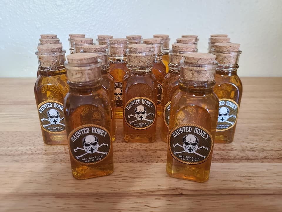 A display of over 10 cork topped jars of golden brown honey with the Haunted Honey label with a masked skull and crossbones from beeguyz.com on a light pine table.