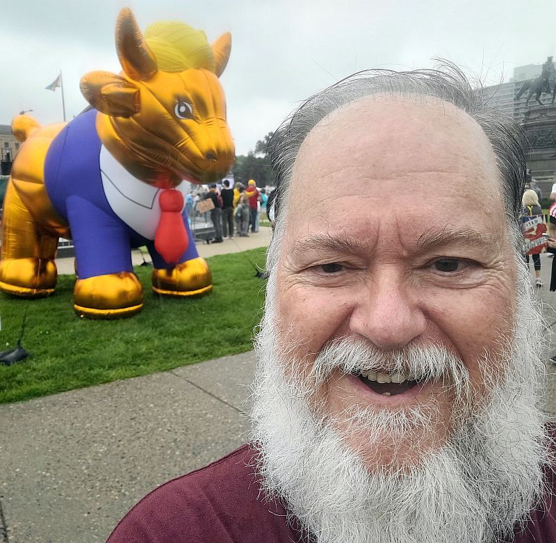 David Milley, a white-bearded man with thinning brown hair laughs in a selfie taken in front of an inflated, angry-looking, gold-colored bull clad in a blue suit jacket and an oversized red tie. A stylized coiffure of pale yellow hair perches uncomfortably between the bull's horns, unmistakably reminiscent of a certain chief executive's hairdo.