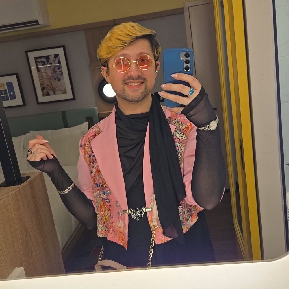 Hotel room mirror selfie. I'm in a long sleeve black mesh shirt under a pink, cropped suit jacket, with lace floral panels. The jacket is held together with a silver rose clasp, the mesh shirt has a long strand that loops around the neck like a scarf. Ive got a watch, silver bracelet, rings, and pink-lensed circular glasses on, and pink and white almond shaped press-on nails
