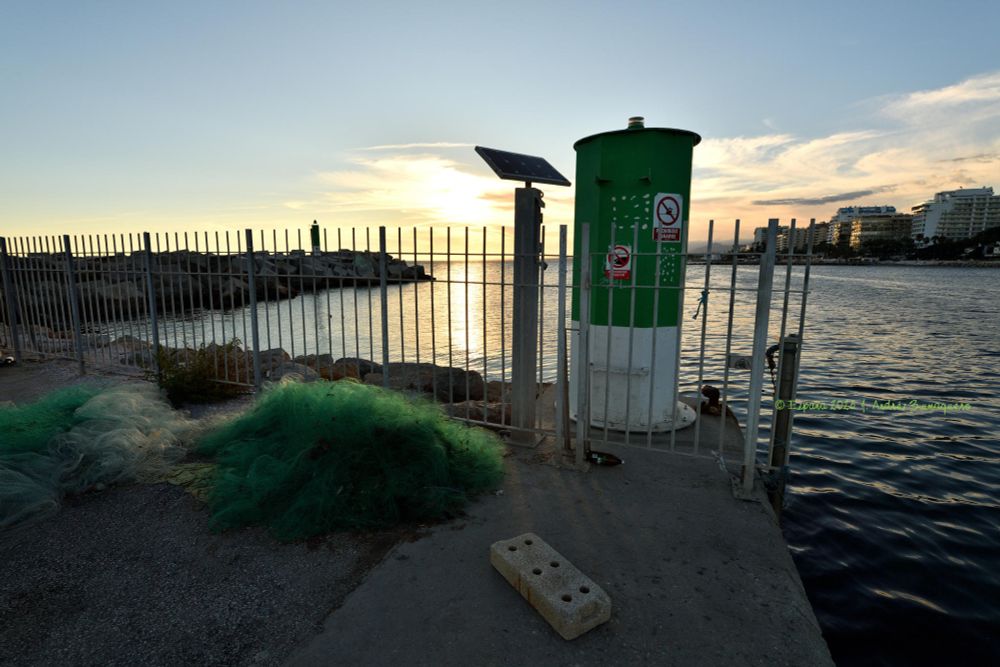 Imagen donde se ve la marca lateral de estribor de entrada al canal del puerto tras una valla. En primer término se ve una malla verde, una red, y un bloque de cemento con seis orificios, posiblemente para fijar postes y vallas. La tarde cae y el cielo tiene alguna nubosidad.