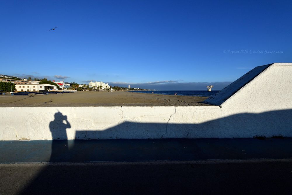 La mitad superior de la fotografía es cielo completamente despejado, donde se aprecia, aunque diminuta, una gaviota. Hay algunas edificaciones, arena de la playa y algo de mar. La parte inferior es un muro blanco que forma parte del muelle de abrigo del puerto, donde se proyecta la sombra del fotógrafo capturando la instantánea.
