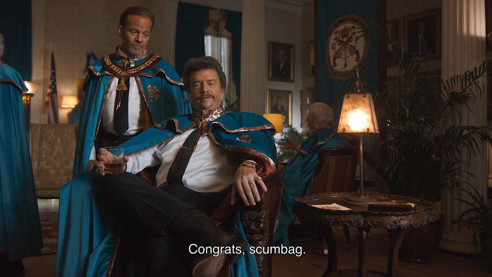 Two men wearing green capes. One is standing and one is sitting