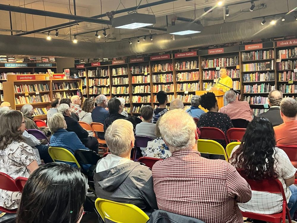 Image from the back of the room with a seated audience listening to Alex Hanna discussing her book "The AI Con"