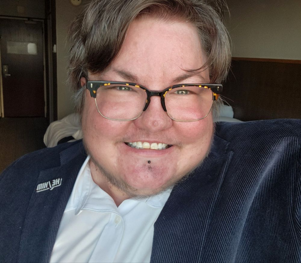 A smiling, fat trans man in a blue blazer and glasses. 