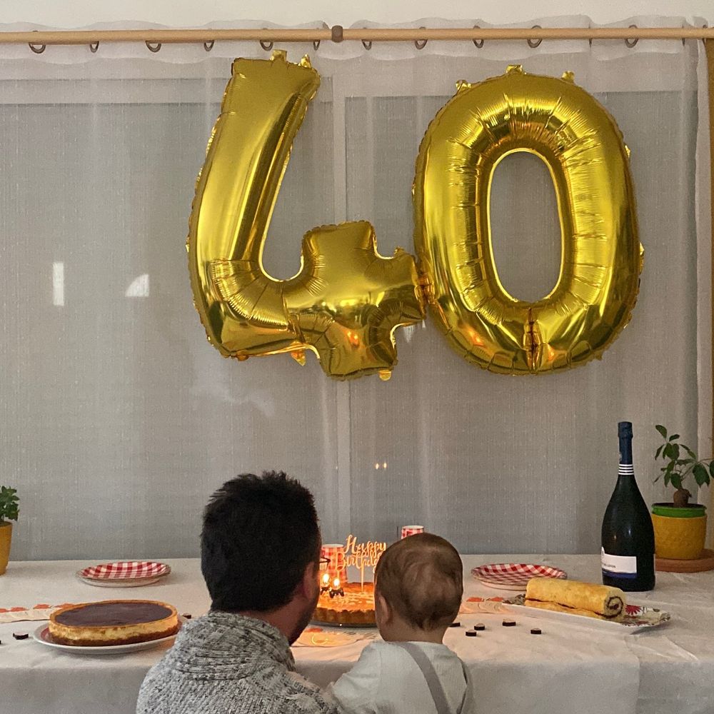 Mon mari (et notre bébé dans ses bras), accroupi et me tournant le dos, prêt à souffler les bougies pour ses 40 ans. Il y a de gros ballons-chiffres dorés (le 4 et le 0) suspendus au-dessus de la table.