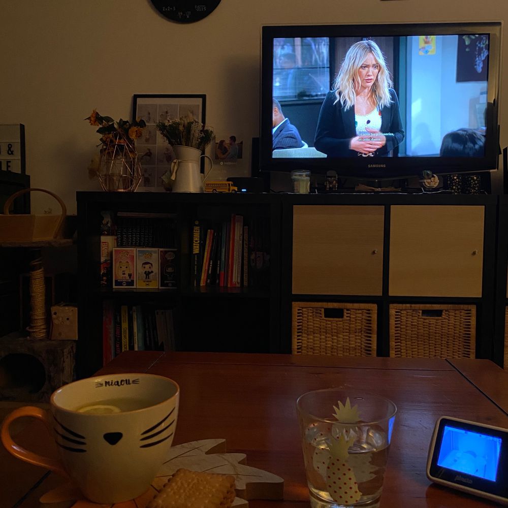 Photo de mon salon avec une infusion de citron, des petits biscuits et le babyphone posés sur ma table basse. Je suis en train de regarder How I met your father donc on voit Hilary Duff à l’écran. La lumière est tamisée.