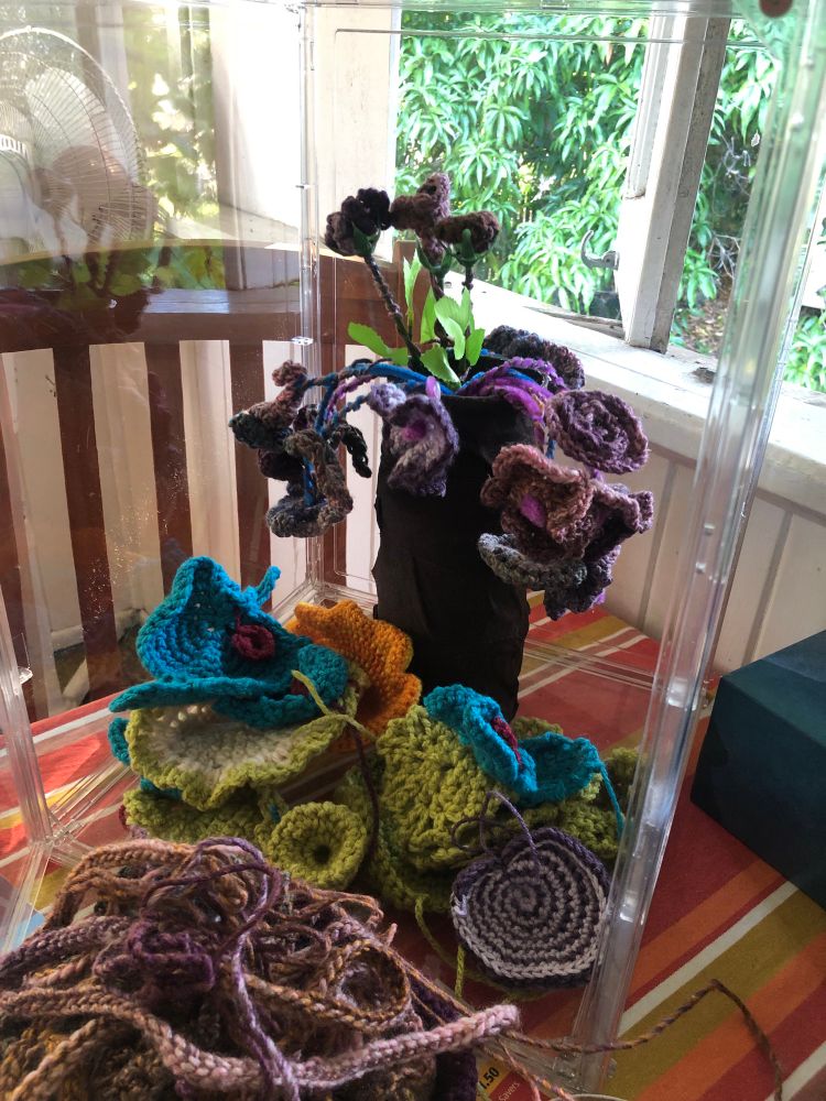 Multiple crocheted flowers in disarray within a Perspex box on a kitchen table. 