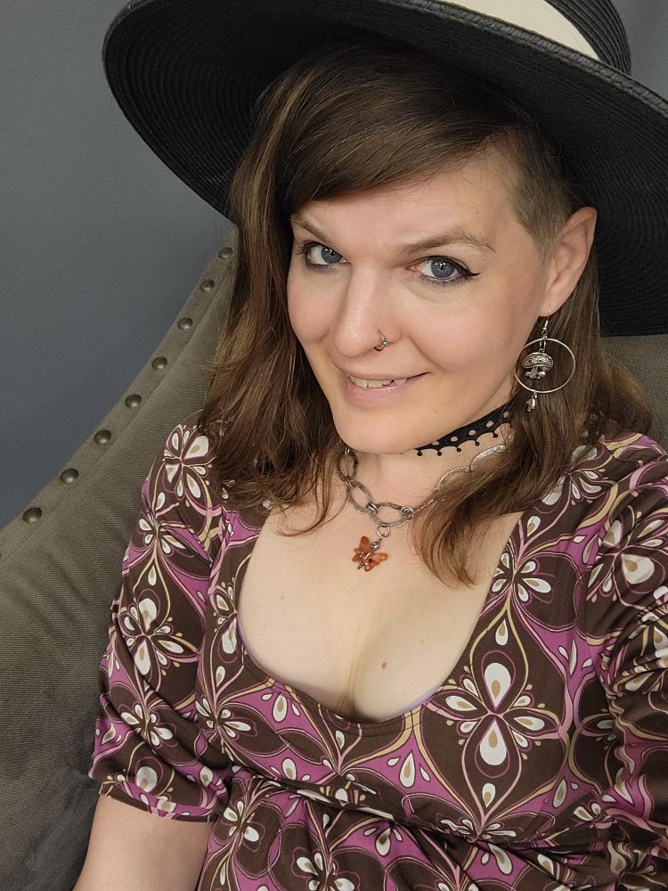 A white woman smiling at a selfie camera with a black hat, slightly alt jewelry and makeup, and a lowish cut, purple and brown dress. 