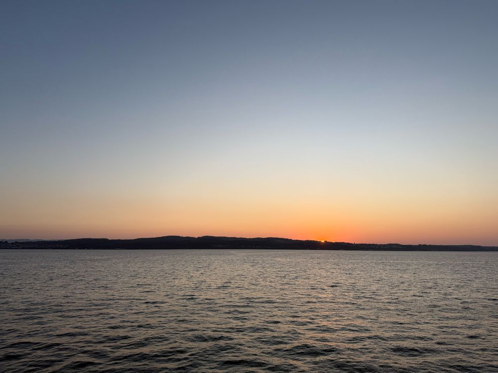 Sonnenaufgang über dem Bodensee, aber man sieht nur einen ganz kleinen Flecken der Sonne über dem Horizont. Der Himmel ist orange und blau und wird im Wasser gespiegelt. 