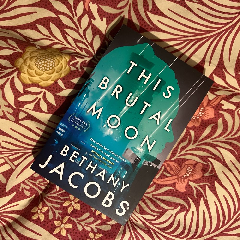 Paperback of This Brutal Moon by Bethany Jacobs, on a red patterned duvet