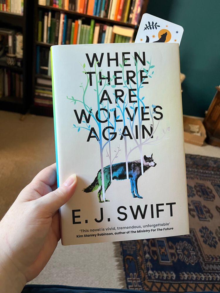 Hardcover of When There Are Wolves Again by E. J. Swift, held in my left hand. There is a bookcase in the background, and a blue, patterned rug.
