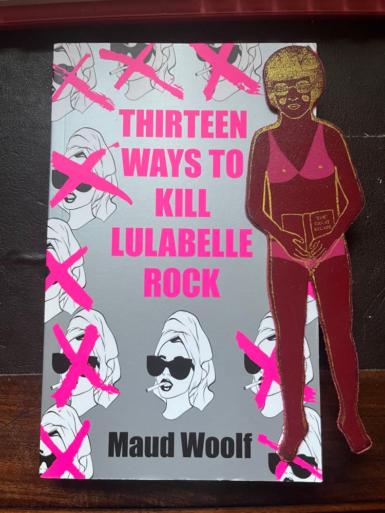 Paperback of Thirteen Ways to Kill Lulabelle Rock by Maud Woolf, which is predominantly grey, but with neon pink text and crosses over white silhouette faces of a woman. A maroon leather bookmark in the shape of a woman with a pink bikini and gold accents sits on top.