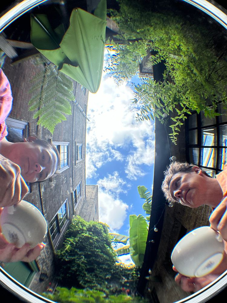 Fish eye photo looking up from a table top, blue with white clouds, visible in the rectangle of sky between tall homes. Plants. Two people drinking coffee. 