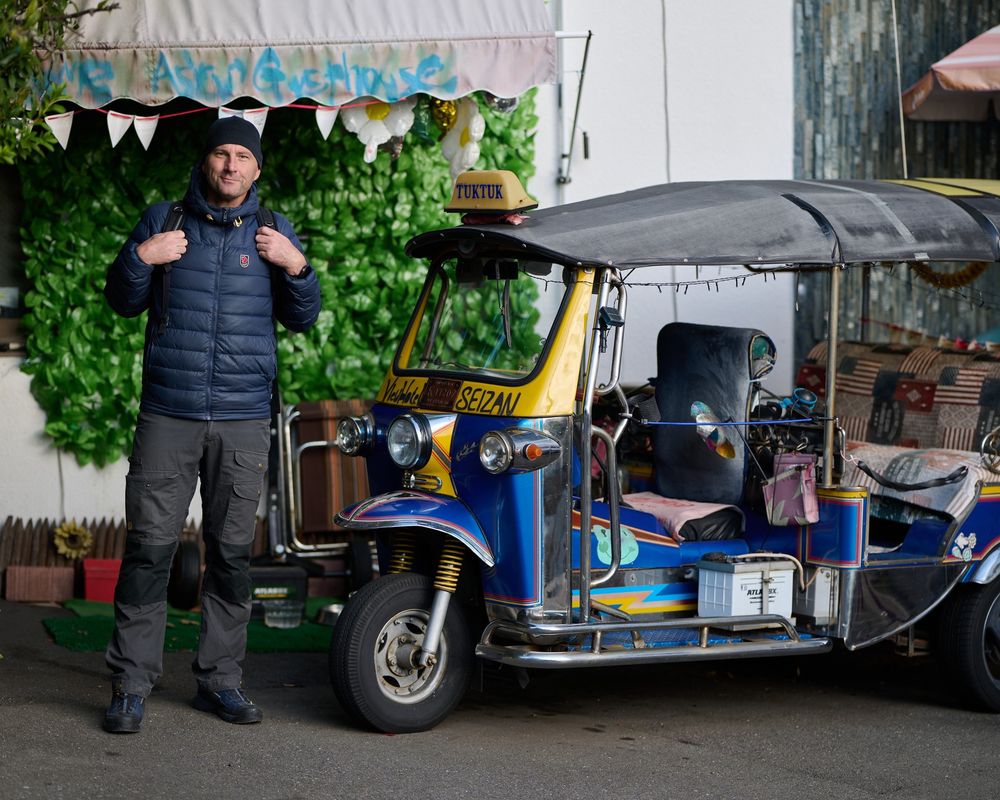 Photo of a handsome man wearing a black toque, blue puffer jacket, and black jeans. He's standing by a small vehicle called a Tuk Tuk. Jeremy has been travelling through Japan the last bit. When he's not travelling the world, he lives in Bangkok, Thailand. 📸 Jeremy Ell 