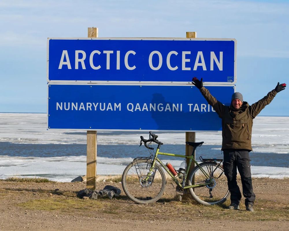 Photo of man wearing a brown jacket and biking pants. He's standing beside a touring bike. The sign says Arctic Ocean. Photo: Jeremy Ell