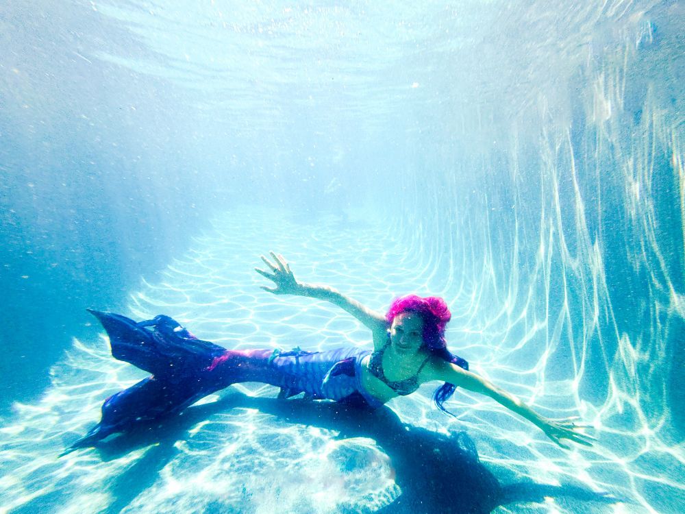 Underwater photo of a mer-person with a dark blue, purple, and pink fish tail, long pink, purple, and blue hair, and a blue snakeskin patterned bikini top. Model is smiling, facing the camera with arms outstretched, the tail is off to the left side of model, while swimming sideways.