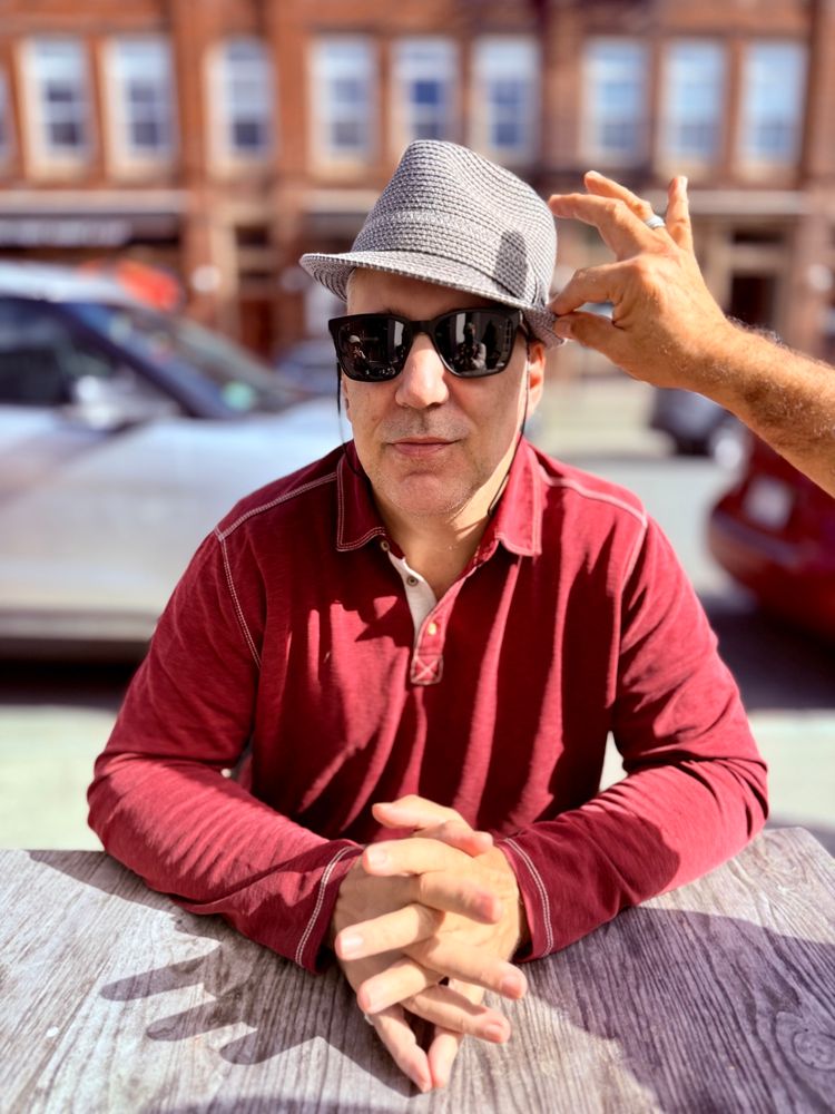 Your correspondent, a white male, wearing a beautiful hat inclined against the sun. A hand comes in snd seems to be adjusting the brim. My sunglasses are also slightly askew, mainly due to my stupid asymmetrical face. 