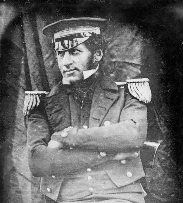 A black and white daguerrotype of Graham Gore, a man in a naval uniform, with his arms crossed and slightly smiling