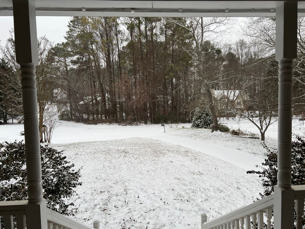 A view from my front porch overlooking my front yard, a portion of my driveway, and cup-de-sac. The road is perfectly covered in snow, and there’s enough snow to cover the grass, but not hide it completely. 