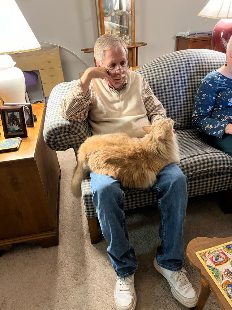 An elderly man with salt colored hair and a mustache is sitting on the couch with a large and slightly fluffy orange cat stretched across his lap. The cat is getting chin scratches and is very pleased with himself. 