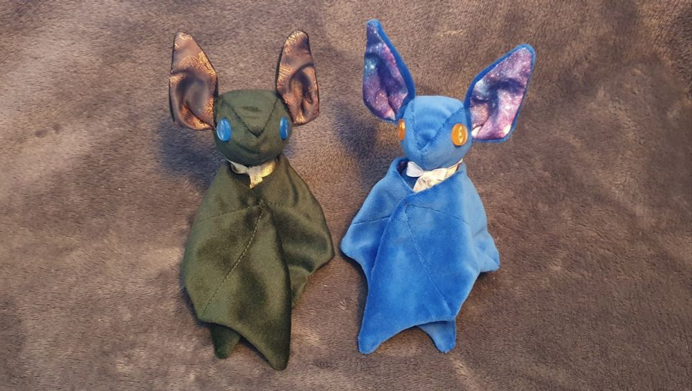 Two bat plushies sat side by side with their wings crossed in front of them. The one of the left is made using dark green velvet, with brown paisley accented fabric for the ears. The one of the right is made using blue soft fabric, with galaxy print fabric for the ears.