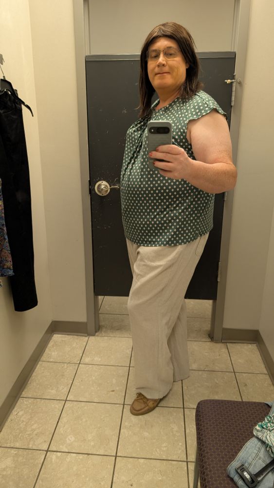 Melissa in a changing room taking a full length selfie.  Wearing a green blouse with a small Chevron like pattern and natural color cotton slacks.  Photo taken turned slightly to the right.