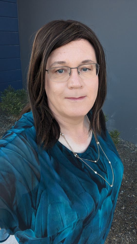 A picture of me, Melissa, wearing a green and black leaf print top.  Smiling and wearing glasses.  Against a gray-blue backdrop with some plants.