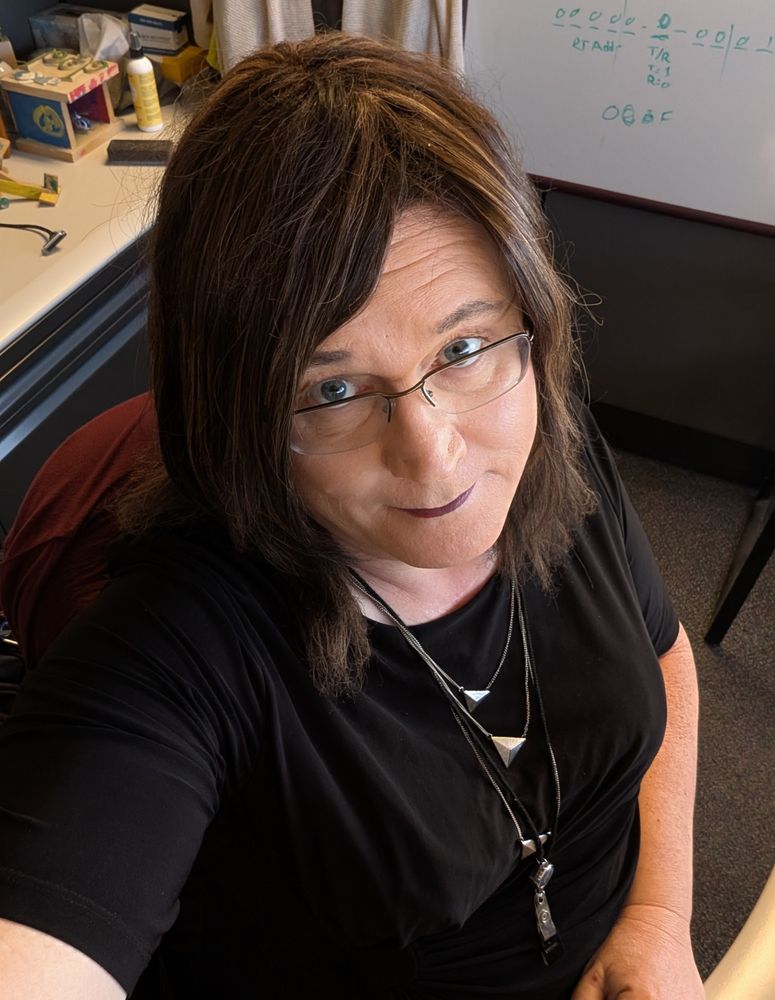 Melissa sitting at her desk, wearing a black dress with a cute chevron necklace.  Wishing it was time to go home for the day. Taken from a high angle.