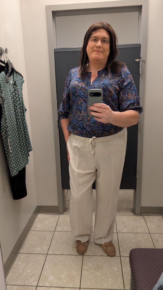 Melissa in a changing room taking a full length selfie.  Wearing a blue, olive, and light red floral print blouse  and natural color cotton slacks.  Photo taken straight on.