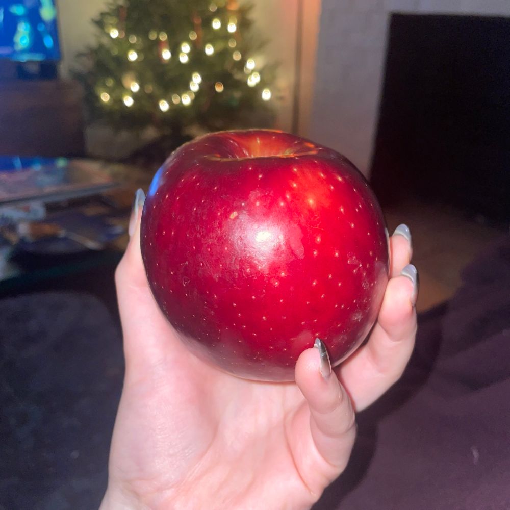 A pale hand holds a bright red and rather large apple. The background is blurry and dimly lighted. 