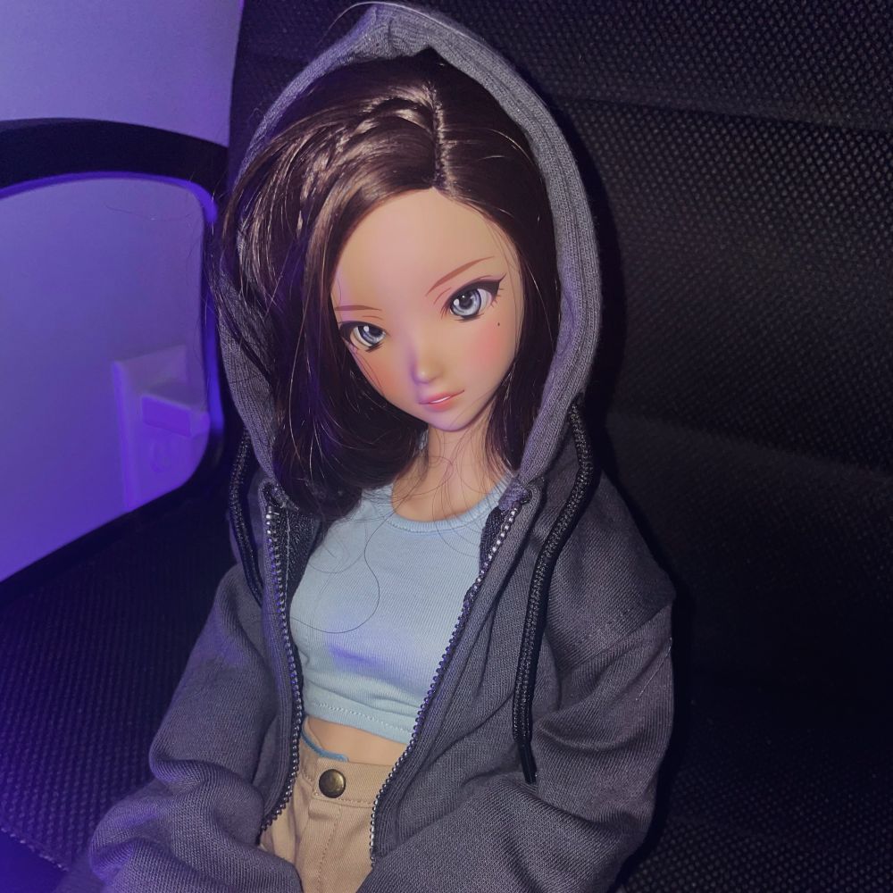 Smart doll never say never in cinnamon with dark brown wavy hair tucked into a grey hooded zip up jacket over a blue top and khaki skirt looking at the camera. The background is black and backlit with a purple LED light 