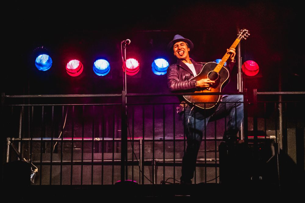Richard performs as Vince in “Woking Phoenix”. He is playing guitar, power posing with one leg up on an amp and his tongue stuck out. He wears blue jeans, a white t-shirt, a purple leather jacket and a purple brimmed hat. In front of him is a black metal railing, and behind him are red and blue stage lights.