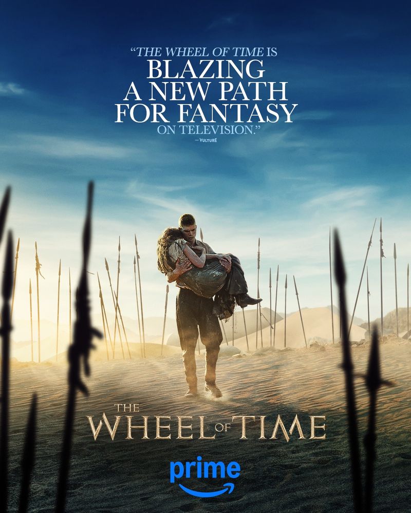 A photo of Rand al'Thor, Josha Stradowski, carrying Moiraine Damodred, Rosamund Pike, through a desert landscape. Text at the top reads “The Wheel of Time is blazing a new path for fantasy on television. – Vulture”. At the bottom there is The Wheel of Time and Prime logo.