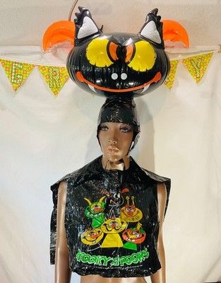 Mannequin in front of blank wall, wearing a 1980s kid's Halloween costume, consisting of a black vinyl smock over the shoulders and a big, vinyl, blow-up head of a bat on its head. The front of the smock is decorated with images of cartoon cats of varying colors and the words "Kooky Spooks" written in neon green at the bottom.