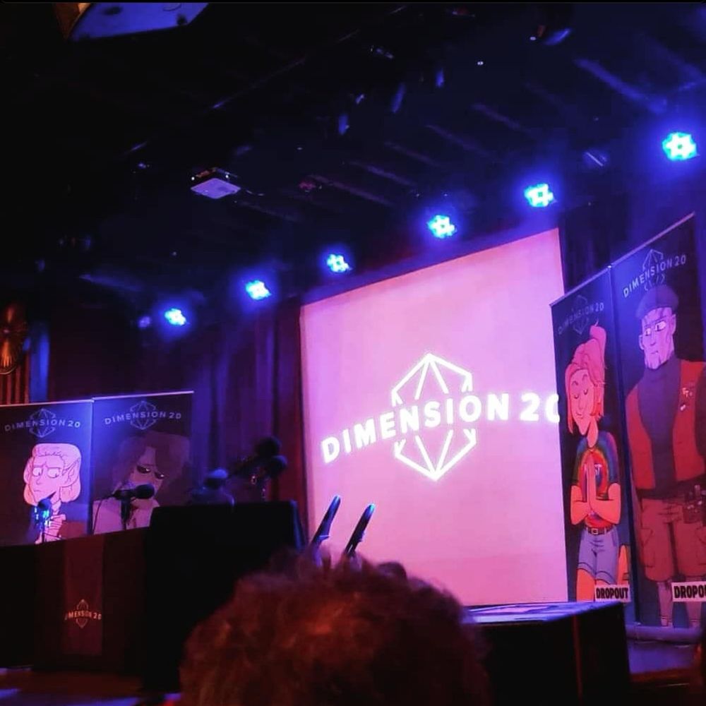 Dimension 20 logo projected on screen. Fantasy High live show at the Bellhouse in 2019. Backdrop of character banners for Adaine, Gorgug, and Kristen. 
