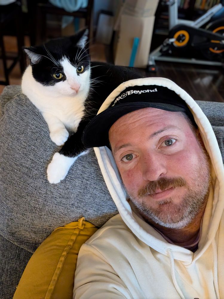 Selfie of me wearing a hoodie settled into the couch while our tuxedo cat, Mascot, looks annoyed while sitting behind me on the back of the couch. 