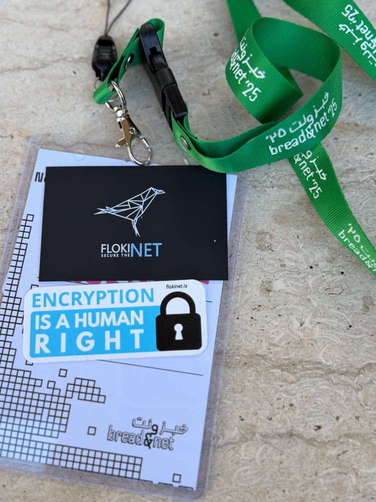 A green Bread&Net 2025 lanyard with a FlokiNET badge and a sticker that reads “Encryption is a human right,” placed on a light stone surface.