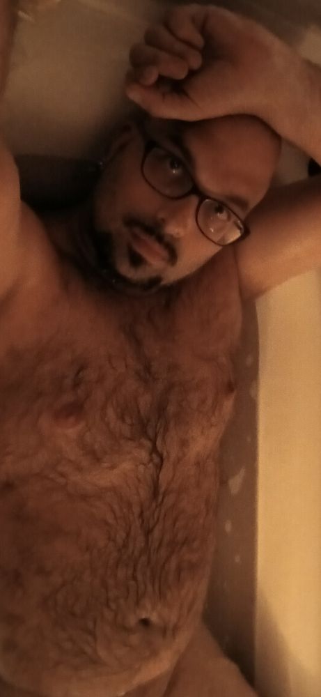 Bald, slightly thick, naked, and hairy gay bear with facial hair framing his jaw and a goatee relaxing in a hot bath in candle light, soap suds, and music. 