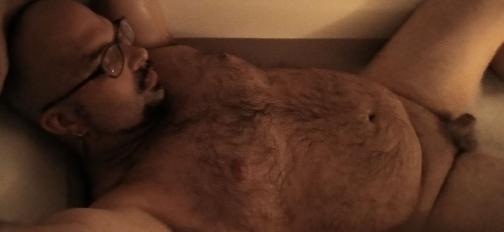 Bald, slightly thick, naked, and hairy gay bear with facial hair framing his jaw and a goatee relaxing in a hot bath in candle light, soap suds, and music. 