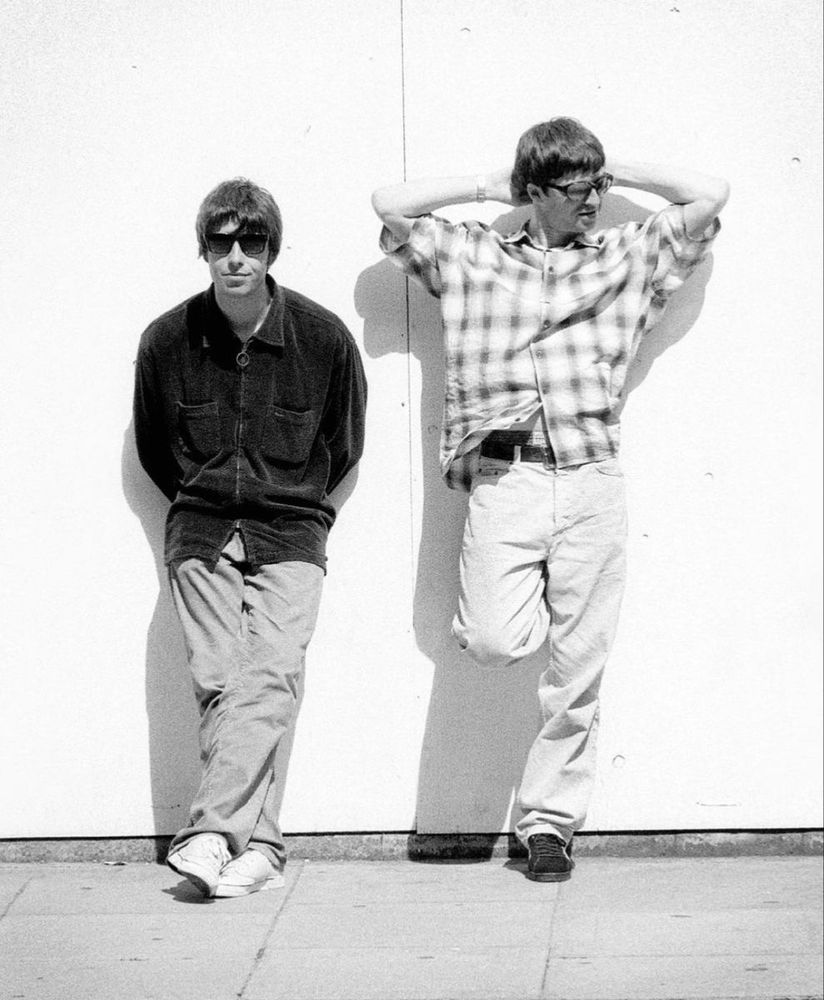 A black and white photo of Liam and Noel Gallagher. Liam is on the left, wearing sunglasses and a dark-colored jacket with his hands behind his back. Noel is to his right, wearing sunglasses and a light-colored gingham button-up with his hands behind his head.