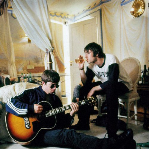 Liam and Noel Gallagher photographed at the Halcyon Hotel. Noel is to the left, sitting on the floor strumming a guitar. Liam is sitting to his right in a chair, drinking a glass of a presumably alcoholic beverage.
