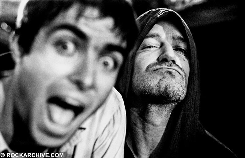A black and white photo of Liam Gallagher and Bono in San Francisco. 1997. Liam is on the left, slightly out of focus. His mouth and eyes are wide open in a smile. His expression takes up half of the photo. Bono is on the right behind Liam, giving the camera a slightly smug expression.