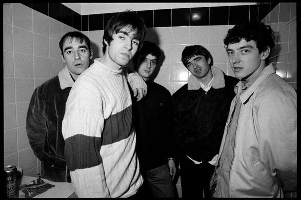 A black and white photo of Oasis, photographed in the cramped “dressing room” at the Jug Of Ale in Moseley. 1994. 
From left to right: Bonehead, Liam, Guigsy, Noel, Tony.

In this photo, Liam has his arm propped up on Guigsy’s shoulder, who is in the back with Noel